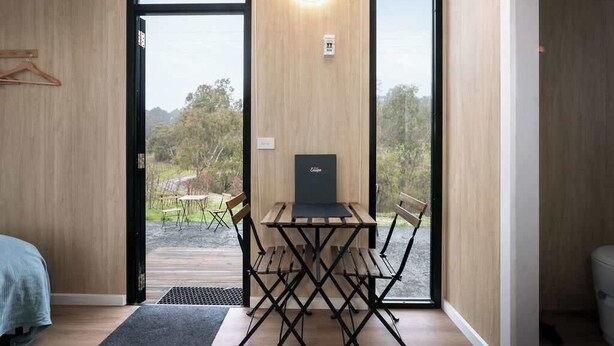 A room with a wooden floor, a bed, a desk with a chair, and a large window with a view of greenery. - Churinga Orchards by Tiny Away (Lenswood)
