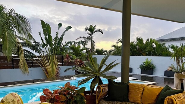 Ein überdachter Außenbereich mit Korbmöbeln, einem Pool und tropischen Pflanzen.