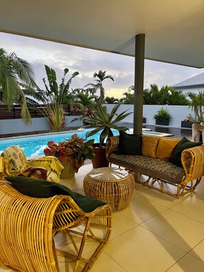 A covered outdoor seating area with wicker furniture, a pool, and tropical plants.