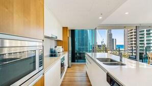 A modern kitchen with stainless steel appliances, wooden cabinets, and a view of tall buildings and the ocean.