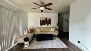 Un salon avec un canapé beige, un pouf foncé, un tapis à motifs, un ventilateur de plafond et une tapisserie murale.