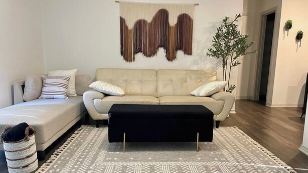 Un salon avec un canapé beige, un pouf noir, un tapis à motifs et une tapisserie murale.