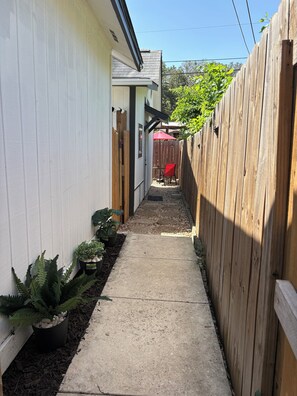 A narrow concrete pathway flanked by a wooden fence and a white building, leading to a patio area with a red umbrella. - Modern & Cozy Studio Near Airport, Downtown & Pearl (San Antonio)