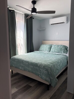 Ein Schlafzimmer mit Bett, Deckenventilator, Klimaanlage und einem Fenster mit Vorhängen.