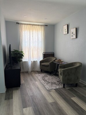 Ein Zimmer mit einem Bett, einem Fernseher, einer Pflanze, einem Sessel, einem Teppich und zwei gerahmten Bildern an der Wand.