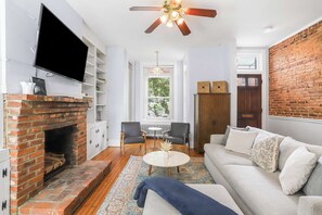 Un salon avec une cheminée en briques, un téléviseur à écran plat, un ventilateur de plafond, un canapé et une table à manger avec des chaises.