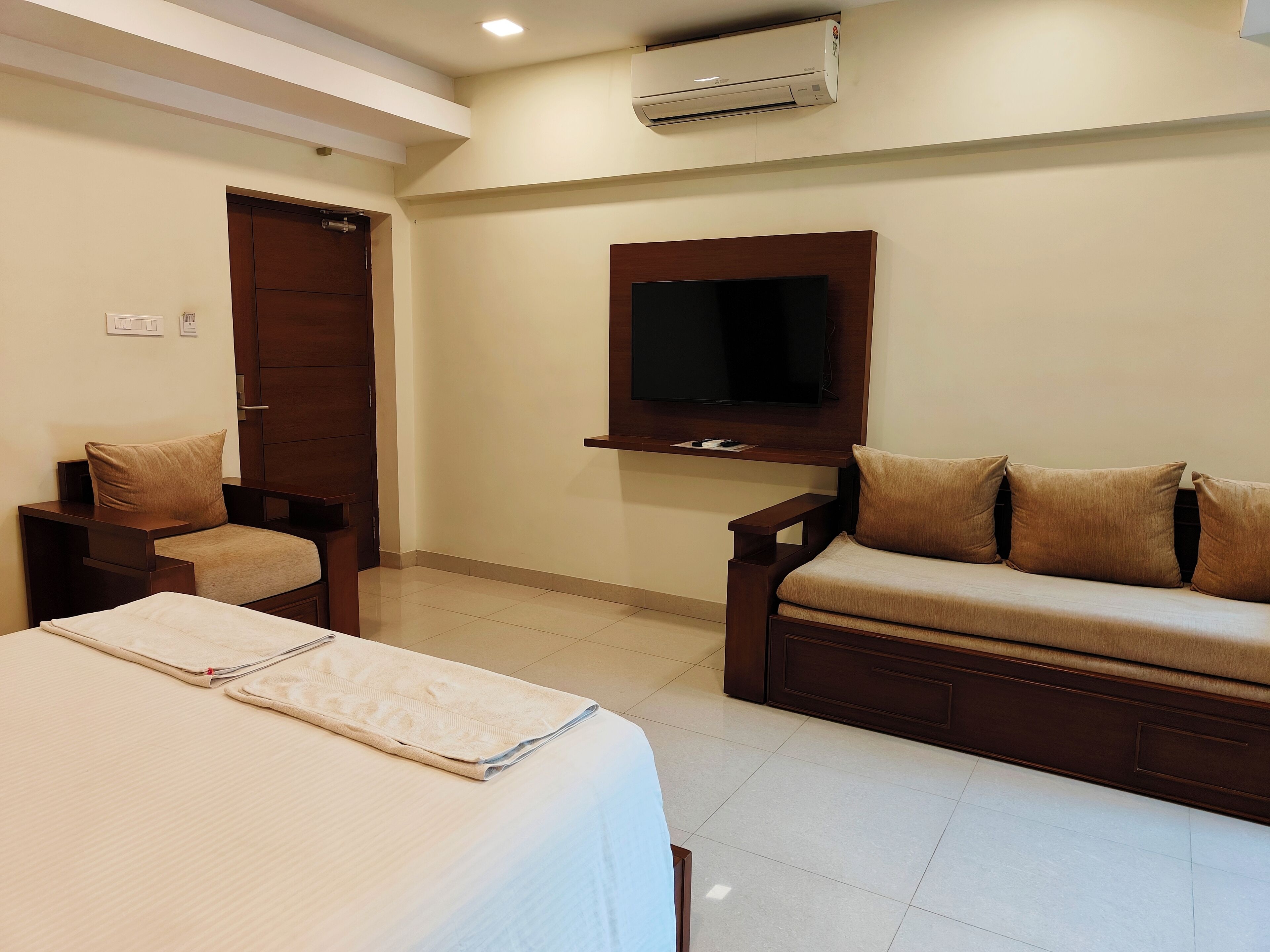 A hotel room with a bed, a sofa, a TV mounted on the wall, and an air conditioning unit.