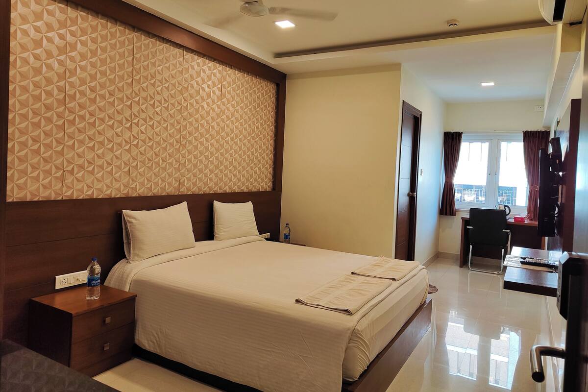 A hotel room with a large bed, a wooden headboard, a nightstand, a desk with a chair, and a window with curtains.