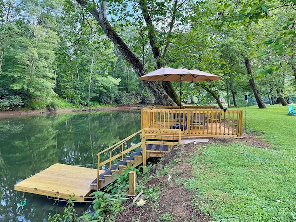 A wooden dock extending into a calm lake, surrounded by lush greenery. - Charming 2 BR, nestled on Buckhannon River, with deck, dock, fire-pit, & kayaks (Volga)