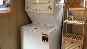 A compact washer and dryer unit with a control panel, a laundry basket on top, and a wooden shelf unit with a detergent bottle.