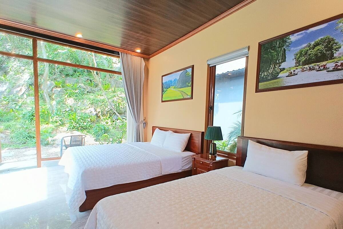A hotel room with two beds, large windows, and a view of greenery outside.