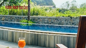 Ein Esstisch mit einem Glas Orangensaft, einer Wassermelone und Toast, mit Blick auf einen Swimmingpool mit einer Steinmauer und Grünflächen im Hintergrund.