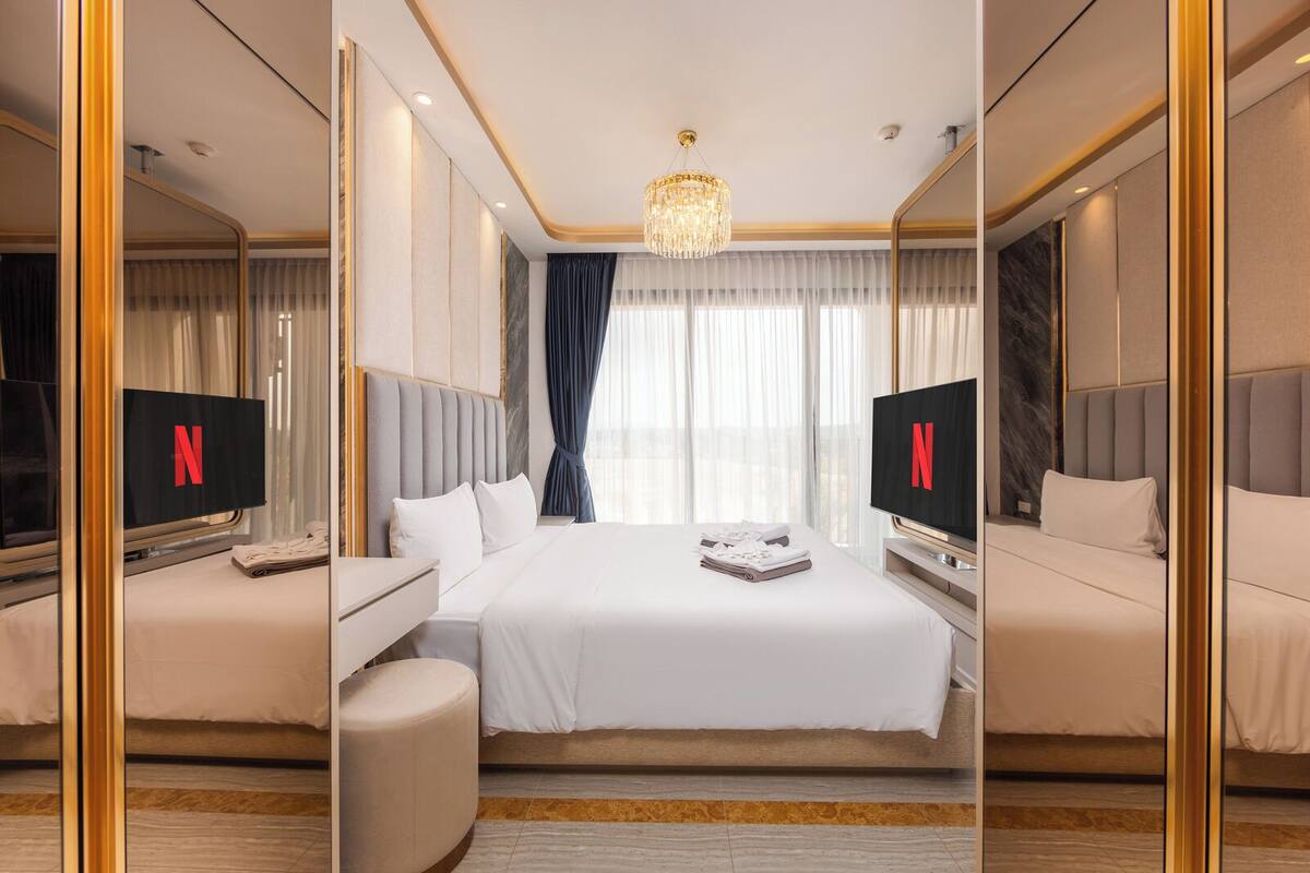 A hotel room with a large bed, two bedside tables, a TV, and a chandelier.