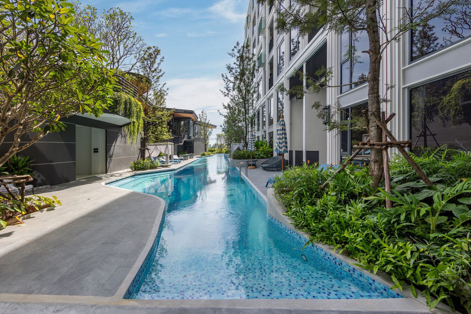 Un espace piscine moderne avec une piscine à l’eau d’un bleu limpide, entourée d’une végétation luxuriante et bordée d’un immeuble à plusieurs étages doté de balcons.