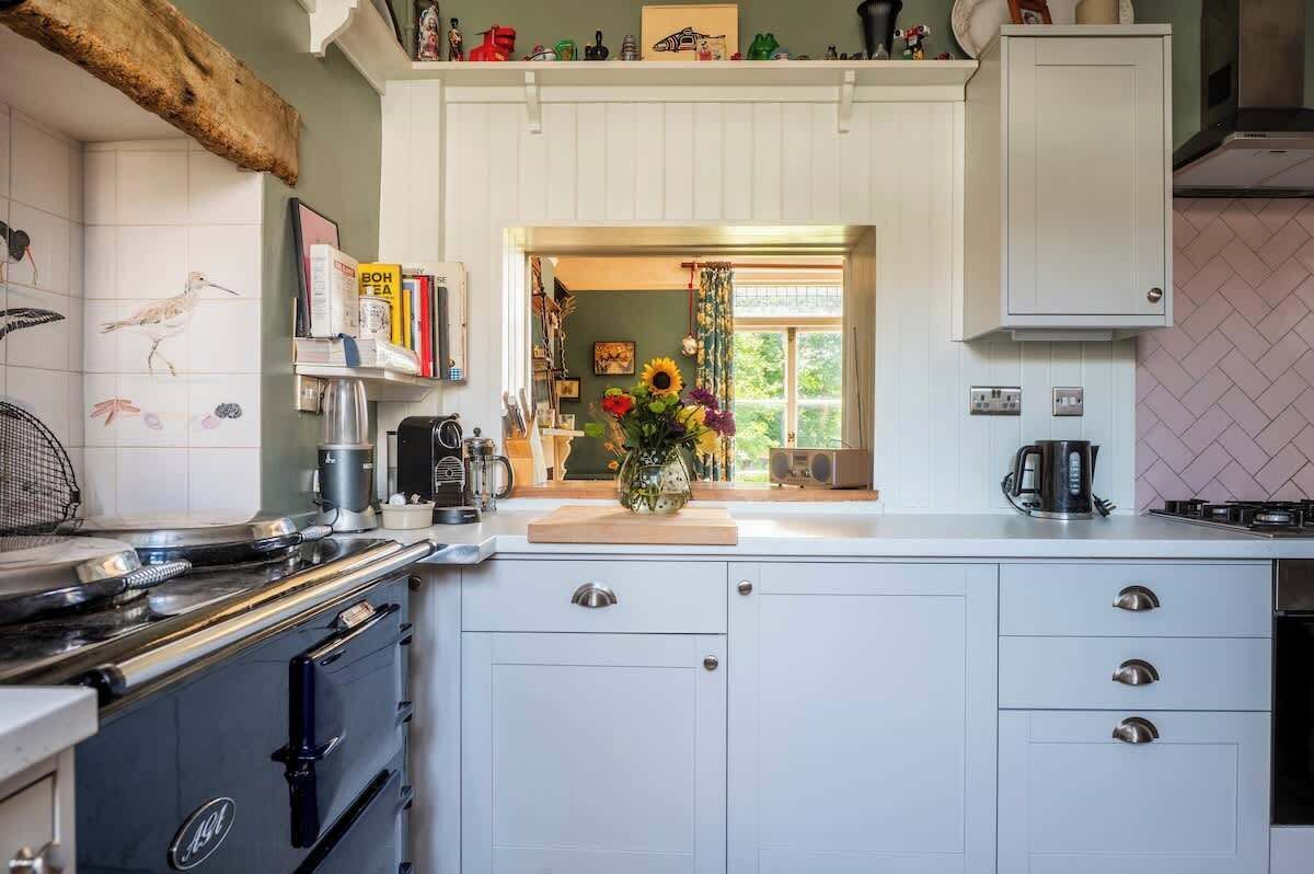 Una cocina con armarios blancos, una estufa y una cafetera. Se ve follaje verde a través de la ventana.