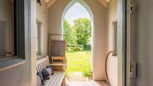 Een veranda met een houten bank en een metalen plank, die uitkomt op een tuin met een bank en een metalen hek.