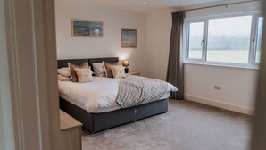A bedroom with a large bed, two bedside tables, a window with curtains, and two wall-mounted paintings. - Modern Country Escape for 12 Guests -Rural Setting (Llanarthney)