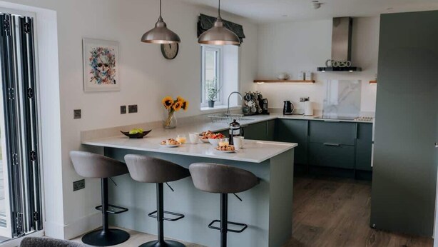 A modern kitchen with a central island, bar stools, pendant lights, and a window providing natural light. - Modern Country Escape for 12 Guests -Rural Setting (Llanarthney)