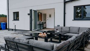 An outdoor patio area with a sofa set, a table with a bottle and glasses, and a view into the house. - Modern Country Escape for 12 Guests -Rural Setting (Llanarthney)