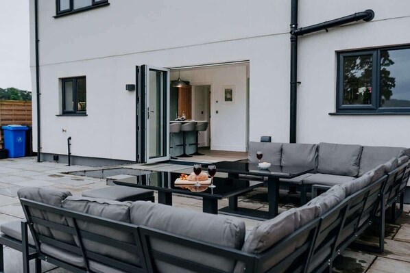 An outdoor patio area with a sofa set, a table with a bottle and glasses, and a view into the house.