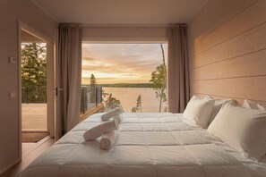 Ett sovrum med en säng, kuddar och utsikt över en sjö i solnedgången.