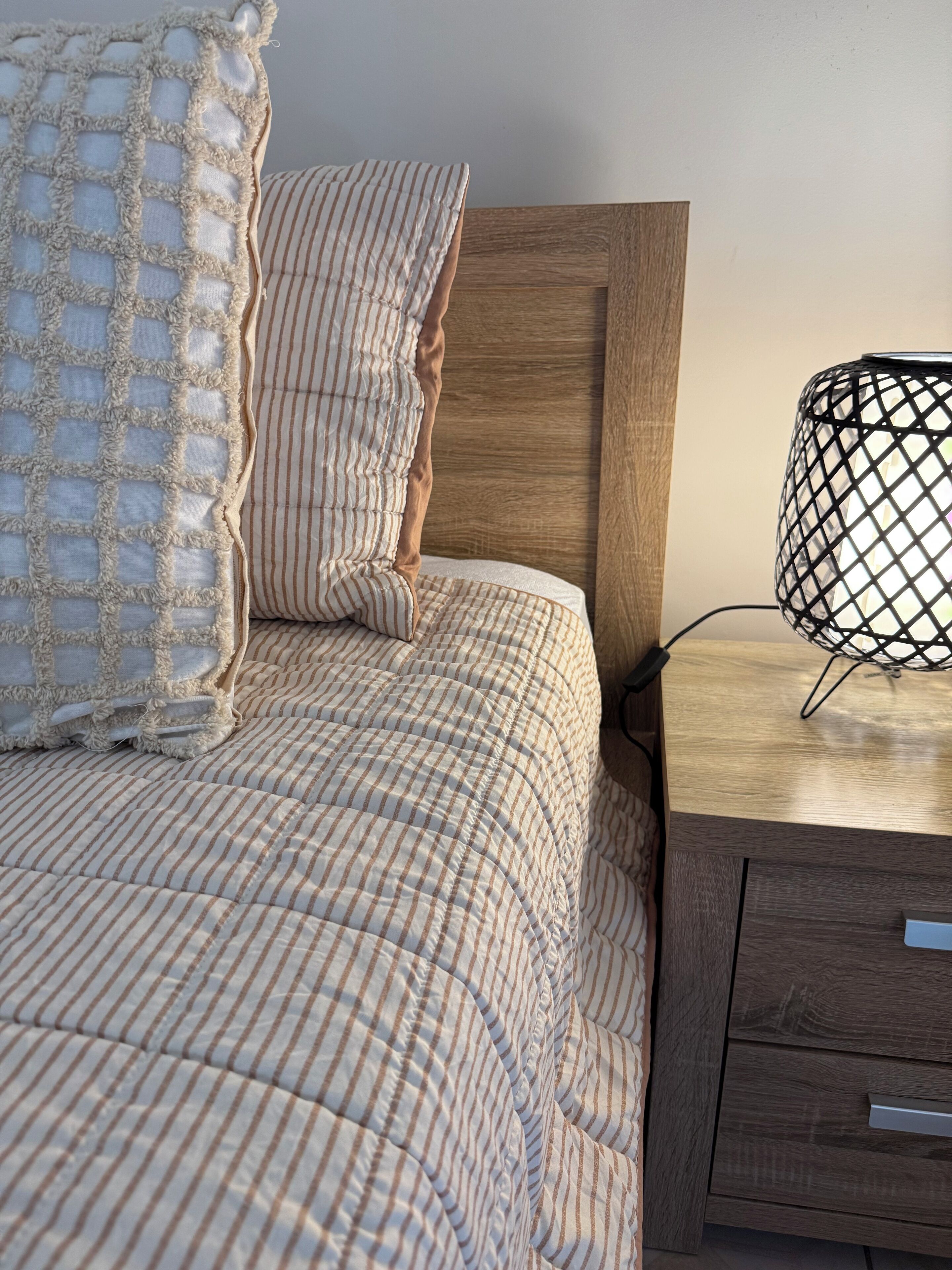 Een net opgemaakt bed met gestreept beddengoed, een houten hoofdbord en een nachtkastje met een lamp.