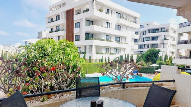 A balcony with a round glass table and two chairs, overlooking a swimming pool and a landscaped garden with various plants and a building in the background. - 1st Class Pool View Apt. (Paphos)