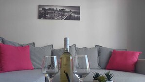A living room with a grey sofa, pink cushions, a table with a wine bottle and two glasses, and a cityscape painting on the wall. - 1st Class Pool View Apt. (Paphos)