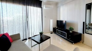 A modern living room with a sofa, a coffee table, a TV on a black stand, and a potted plant. - 1st Class Pool View Apt. (Paphos)