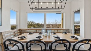 Un comedor con una mesa grande, cuatro sillas, una araña y ventanas amplias con vistas al exterior.