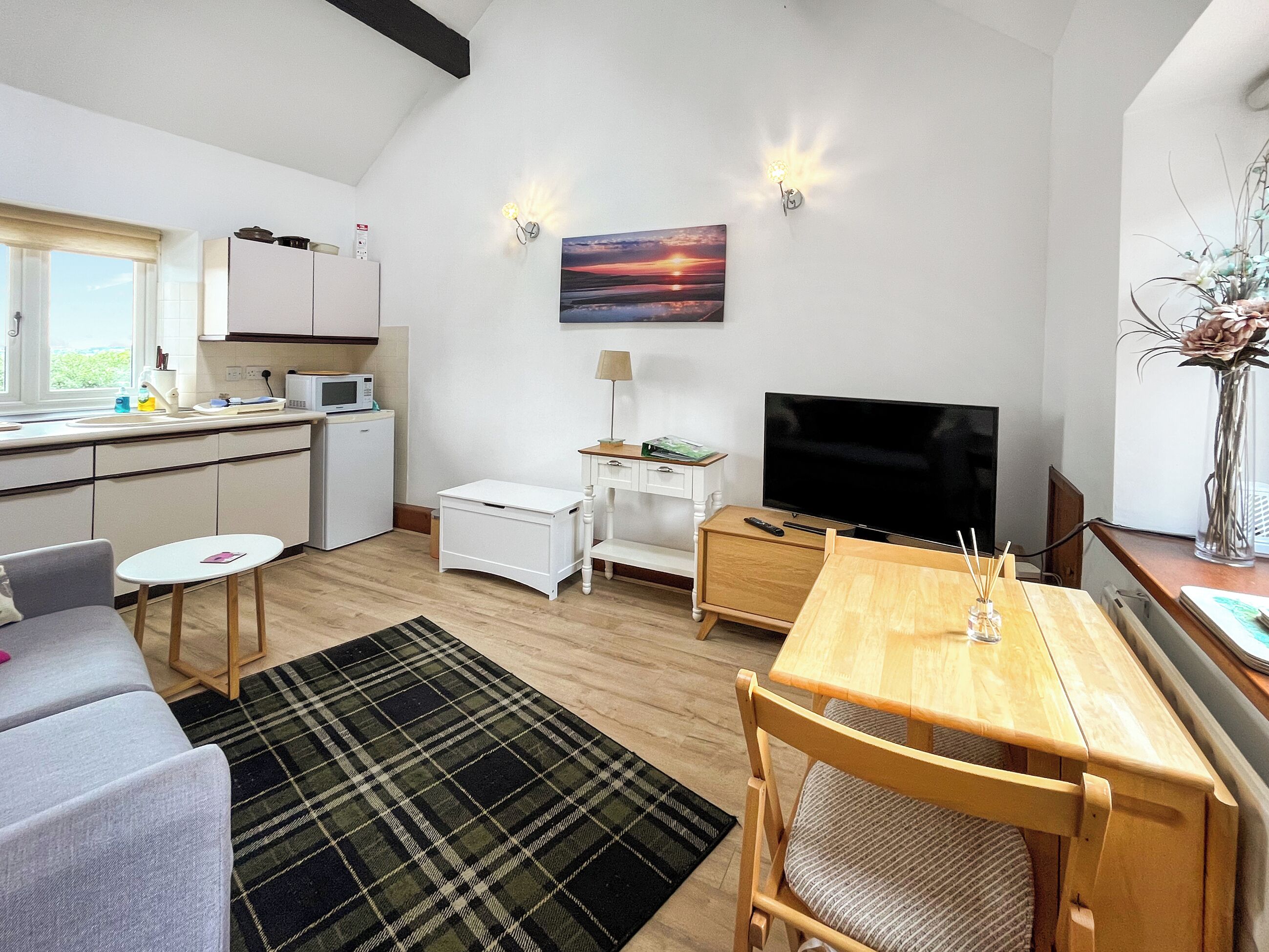 A modern kitchen with white cabinets, a microwave, and a small refrigerator. There is a dining area with a wooden table and chairs, a TV, and a painting on the wall.