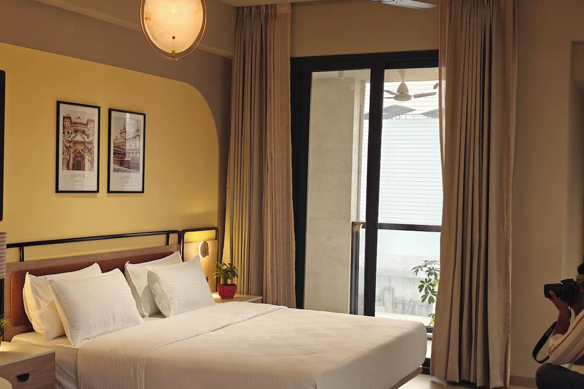 A hotel room with a large bed, a nightstand, a lamp, a framed picture of Jaipur, and a window with curtains.