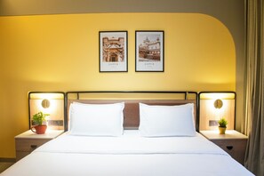 Habitación de hotel con una cama, dos mesitas de noche con lámparas, una planta en maceta y dos cuadros en la pared.
