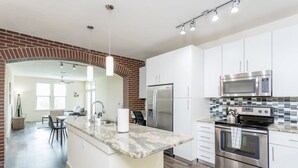 Eine moderne Küche mit weißen Schränken, einem Edelstahlkühlschrank, einer Mikrowelle und einem Backofen. Eine große Insel mit Granitplatte und einer Pendelleuchte.