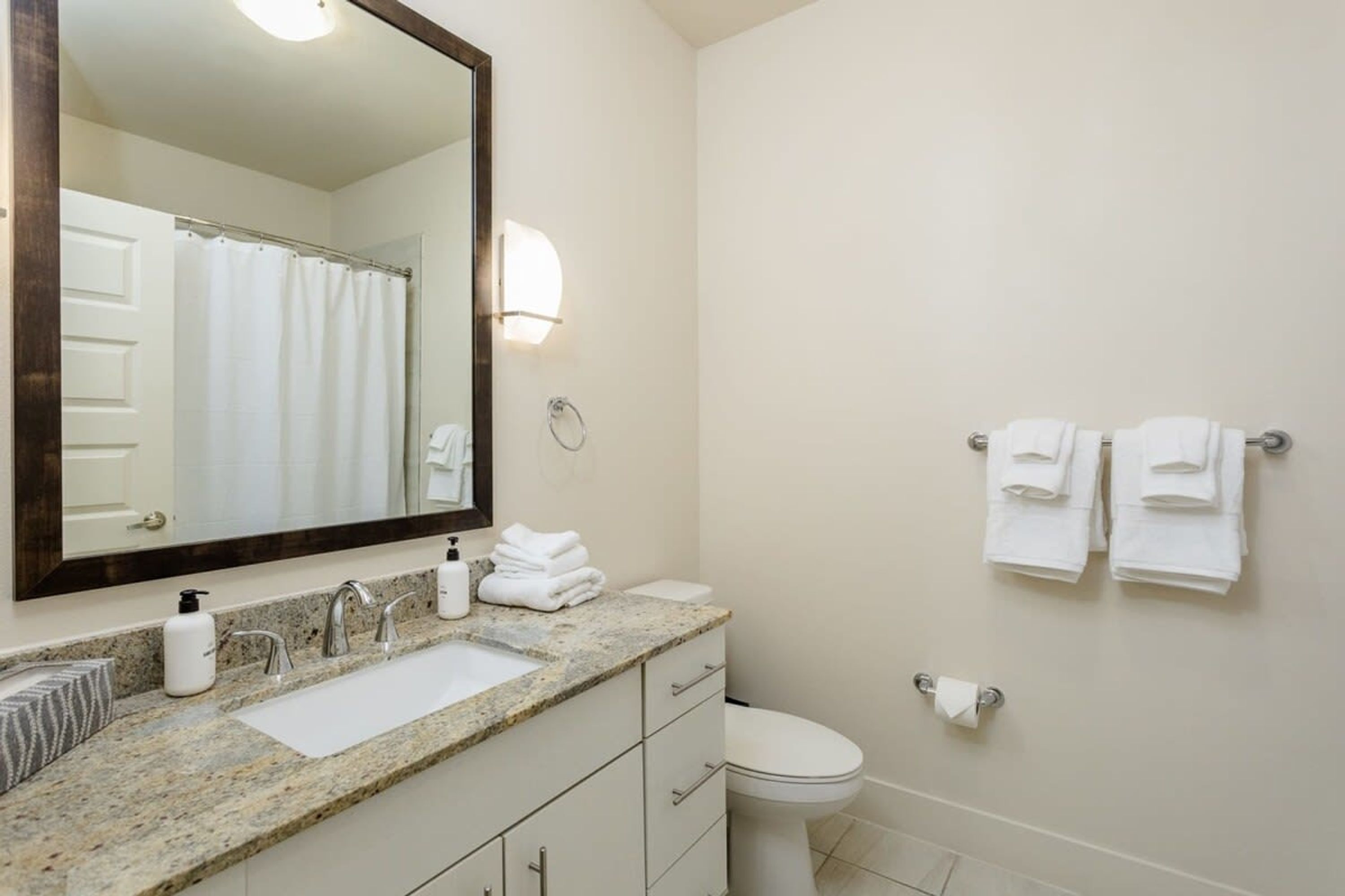A bathroom with a granite countertop, a white sink, a mirror, a toilet, and a towel rack with folded towels.