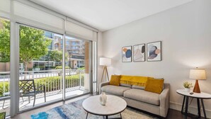 A modern living room with a sofa, a coffee table, and a lamp. There are large windows with a view of buildings and greenery outside. - The Preston Charm I Premium 1BR in Dallas (Dallas)