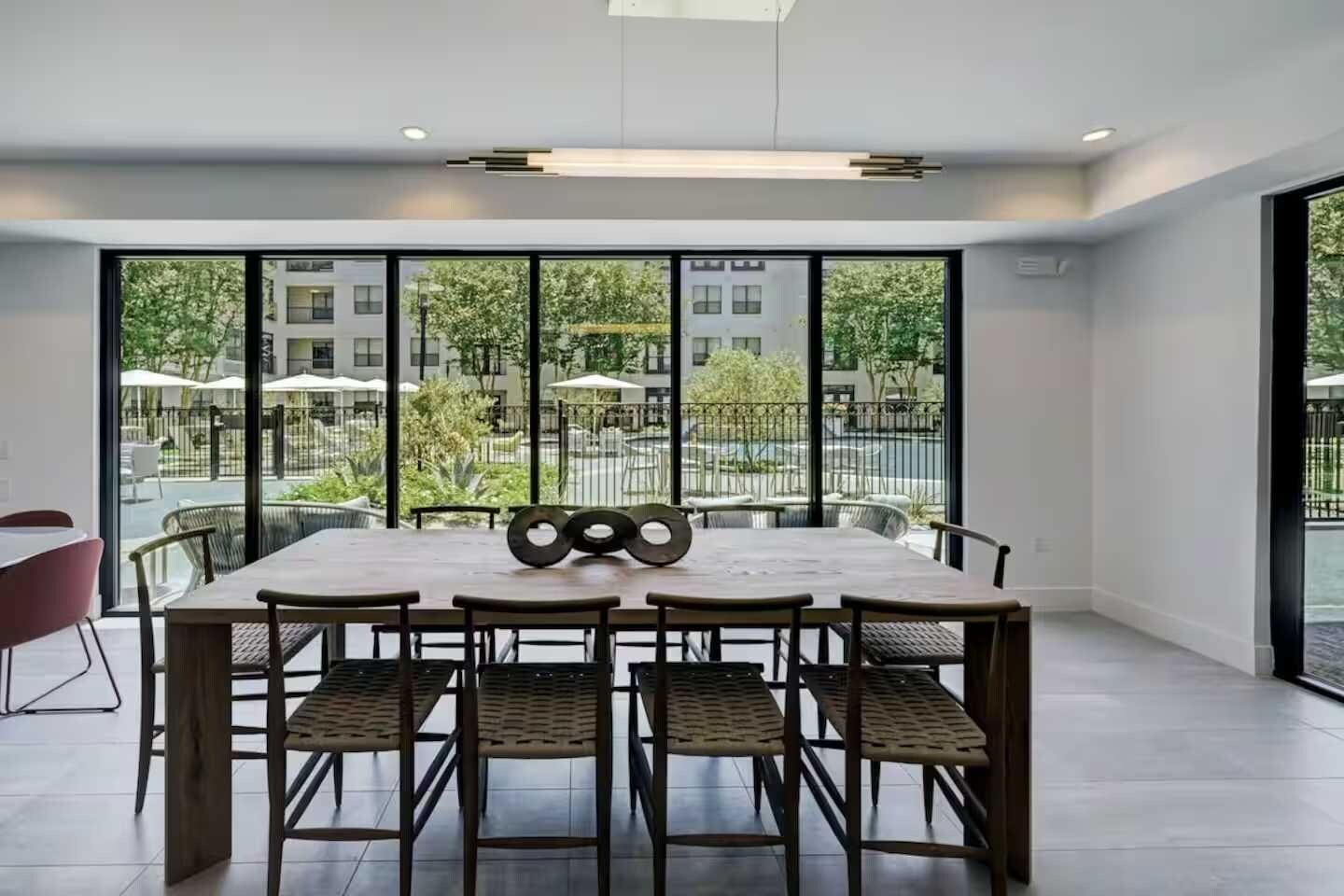 A modern dining area with a large table and chairs, large windows offering a view of the outdoors, and a minimalist ceiling design.