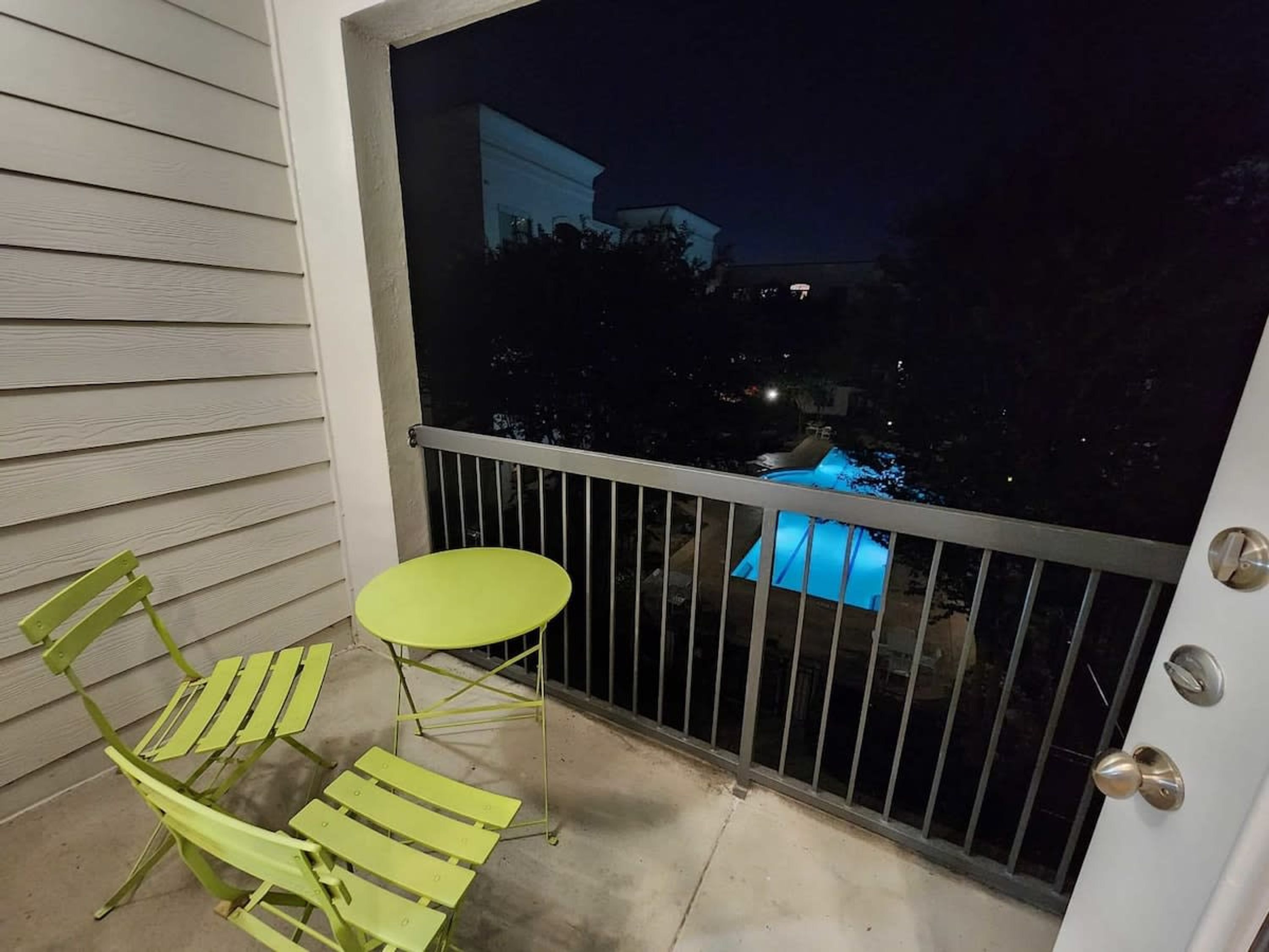 En balkong med en grön stol och ett bord, en pool med blått ljus och utsikt över natthimlen.