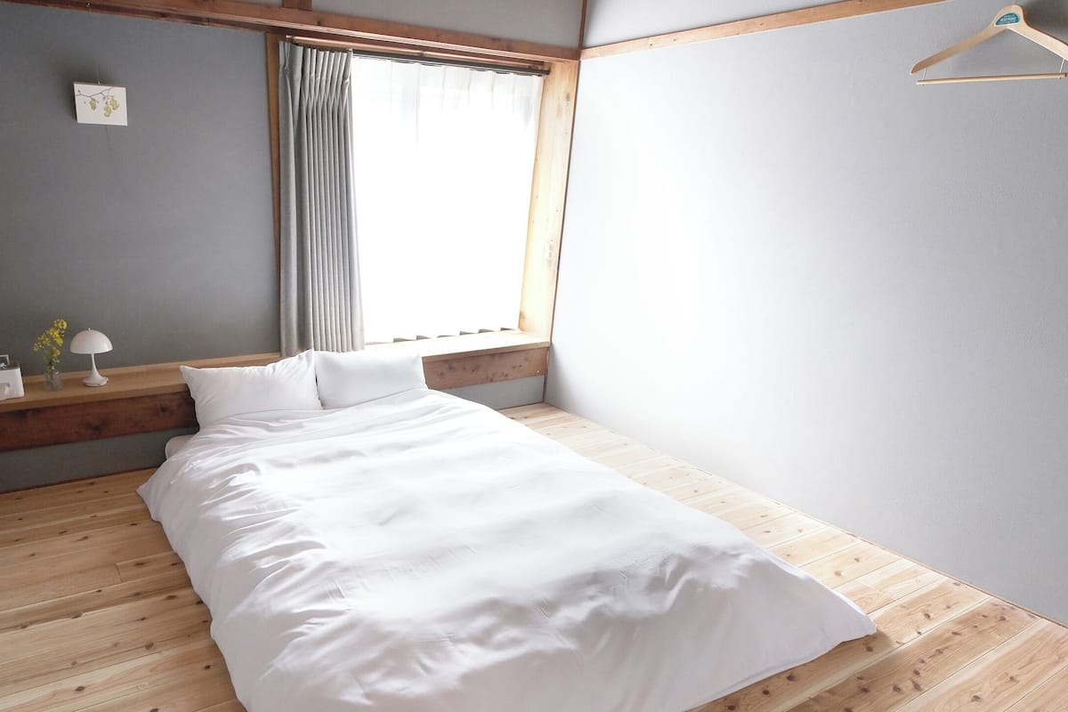 Ein Schlafzimmer mit Holzboden, einem Bett mit weißer Bettwäsche, einem Nachttisch mit Lampe und einem Fenster mit Vorhängen.