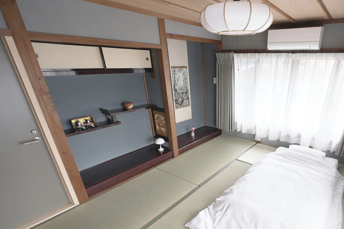 Ein Zimmer mit Tatami-Boden, einem Bett, einem Schreibtisch mit Lampe, einem Regal mit Dekoration und einem Fenster mit Vorhängen.