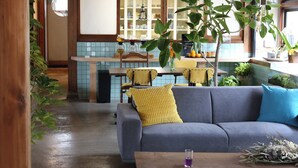 A cozy living room with a gray sofa, a wooden coffee table, and a yellow cushion. A kitchen with blue tiles and wooden cabinets is visible in the background. - junos (Nahari)