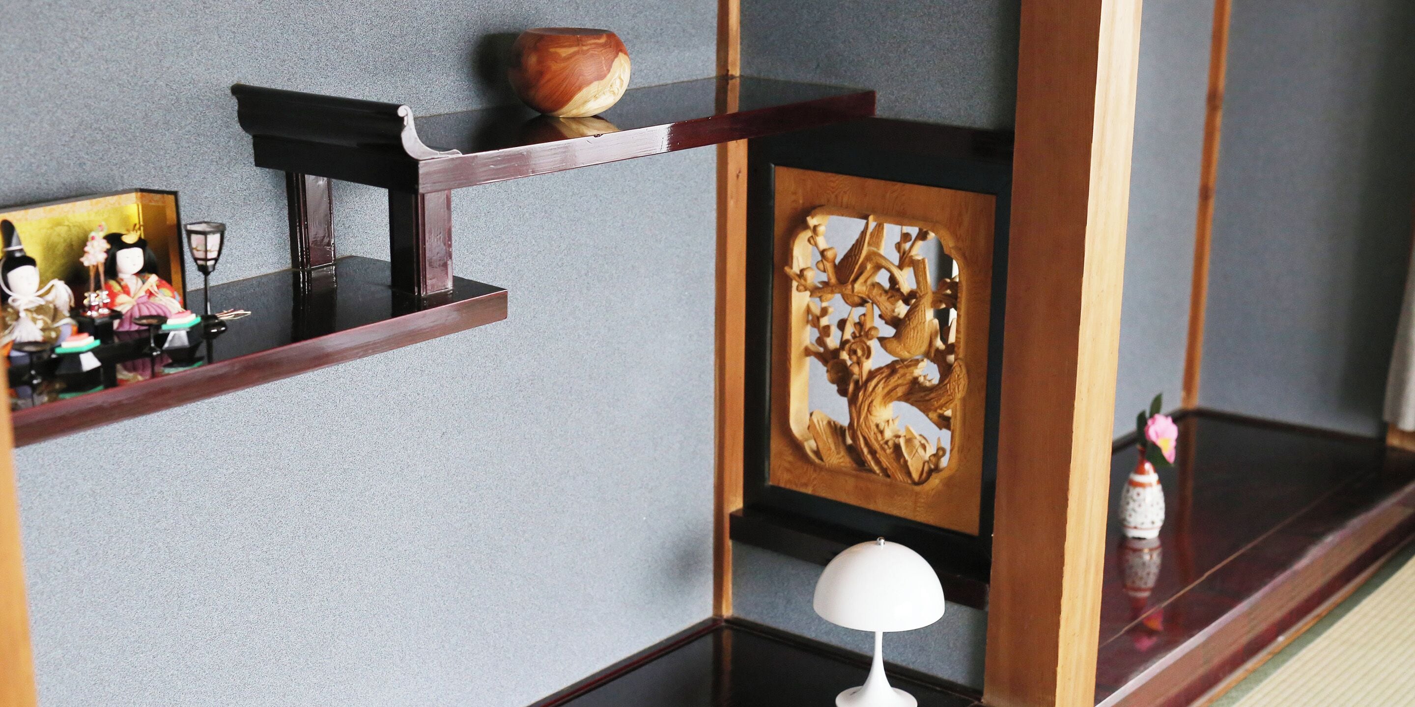 A corner of a room with a wooden shelf holding decorative items, a wall with a carved wooden panel, and a small white lamp.