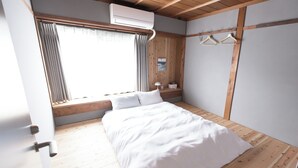 A bedroom with a wooden floor, a bed with white bedding, an air conditioning unit, a window with curtains, and wooden wall paneling. - junos (Nahari)