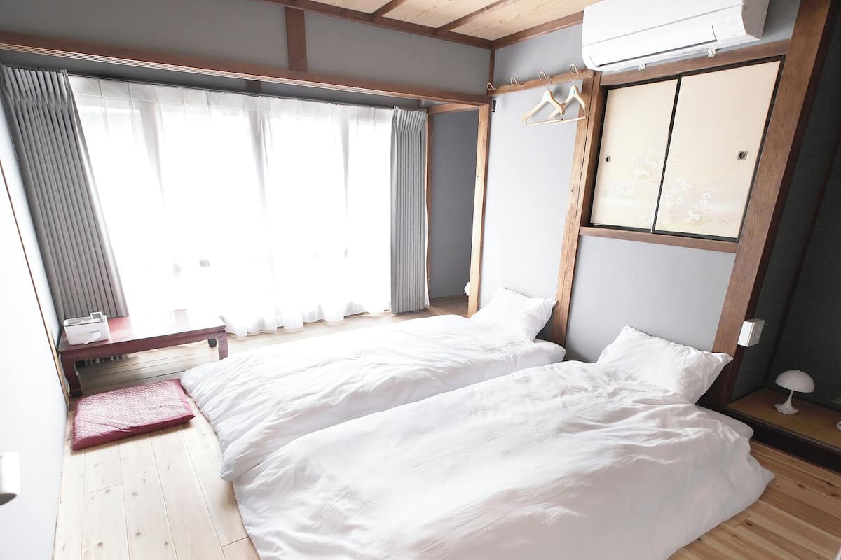 Ein Zimmer mit zwei Betten, Holzboden, einem Fenster mit Vorhängen, einem Nachttisch mit Lampe und einer Wand-Klimaanlage.