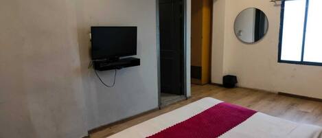 A bedroom with a bed, a TV mounted on the wall, a door leading to another room, and a window with blinds.
