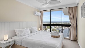 A hotel room with a large bed, a nightstand with a lamp, a ceiling fan, and a view of a cityscape through large windows. - Luxury Beachfront 1BR Apt with Stunning Ocean View in the centre of Surfers (Surfers Paradise)