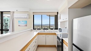 A modern kitchen with white cabinets, a stainless steel refrigerator, a microwave, and a view of a cityscape through large windows. - Luxury Beachfront 1BR Apt with Stunning Ocean View in the centre of Surfers (Surfers Paradise)