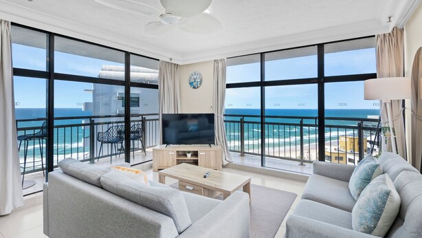 A modern living room with a large TV, grey sofas, and a balcony with ocean views. - Luxury Beachfront 1BR Apt with Stunning Ocean View in the centre of Surfers (Surfers Paradise)