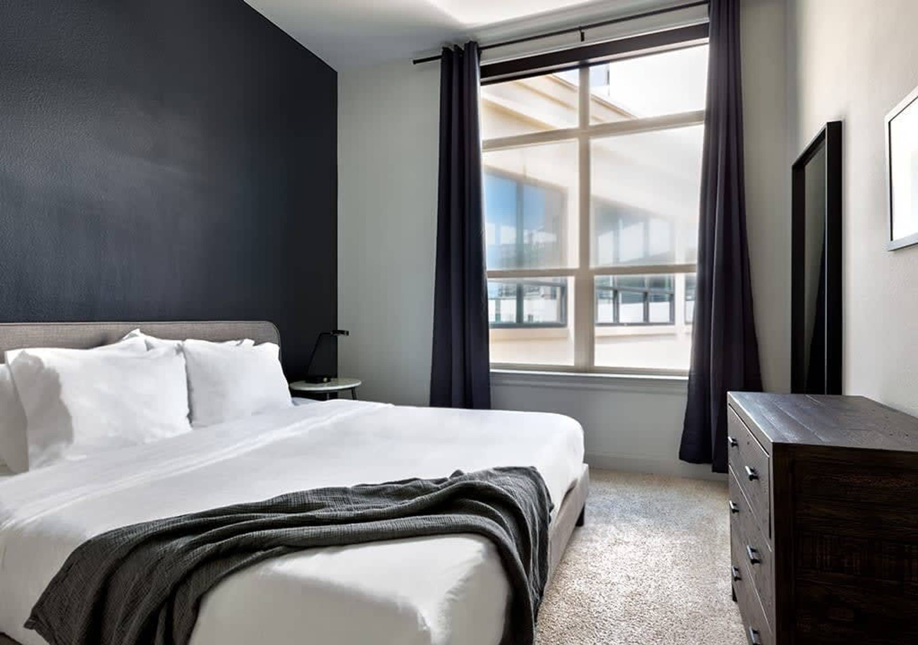 Ein Hotelzimmer mit einem großen Bett, einem Schminktisch, einem Nachttisch und einem Fenster mit Vorhängen.