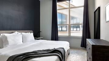 Ein Hotelzimmer mit einem großen Bett, einem Schminktisch, einem Nachttisch und einem Fenster mit Vorhängen.